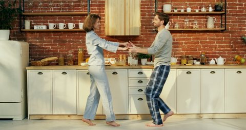 Young happy Caucasian just-married couple in pajamas dancing nice together in the cozy kitchen early in the morning.