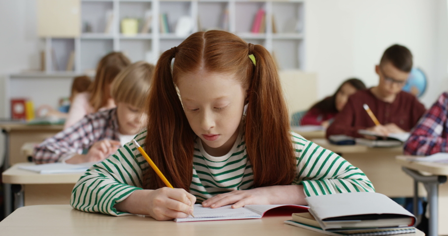 Portrait shot of the Caucasian teen red-haired schoolgirl writing in the copybook at the lesson in the classroom and smiling cheerfully to the camera.