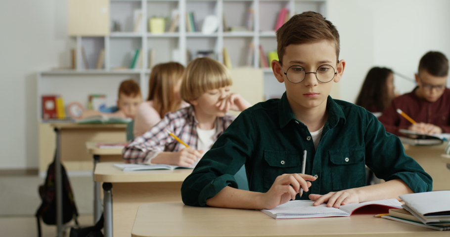 Caucasian teen boy in glasses writing a test in the copybook and thinking over the right answer at the control exam at school.