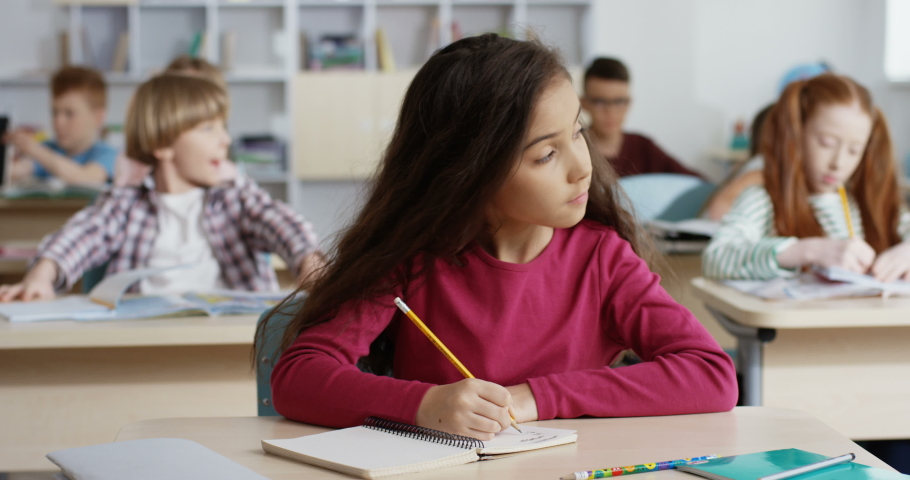 Small beautiful schoolgirl with long dark hair wriying with a pencil an exercise in the copybook during control test at the school lesson.