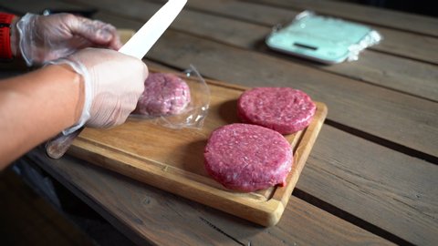 Man in gloves and with knife opening raw burgers in the kitchen