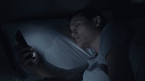 Close-up pan from mobile phone to a young man's face as he lies in bed at night using his mobile phone. Concept: Poor sleep health