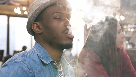 Portrait shot of african young man smoking hookah in the bar with young attractive woman on the background.