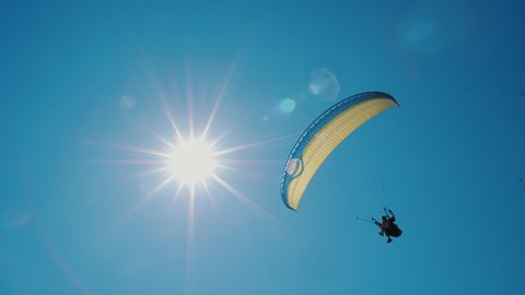 Extreme paraglider flying against a clear blue sky, sunbeam shines into camera. Man with instructor flies with parachute. 4k, slow motion.