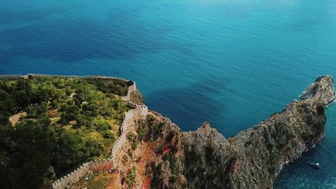 Aerial view around ruins and ancient historic Alanya fortress walls on rocky hills and riffs at background of azure mediterranean sea with sailing ship. Antalya Province, Turkey, 4k footage.