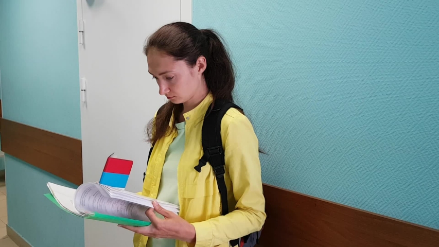 Brunette girl in the room of one of the institutions holding documents in her hands, flipping through the paper and checks some data. Waiting her turn