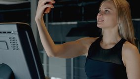 Close up and profile side view of girl, wearing in trendy sport wear training on bicycle machine inside sport center with modern interior. She using cellular phone to speaking on video call