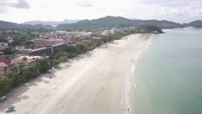 Aerial drone shot for Chenang Beach in Langkawi Island in Malaysia during the morning and people on the beach, with the sun lights and cloudy day, and houses and resorts surrounding.