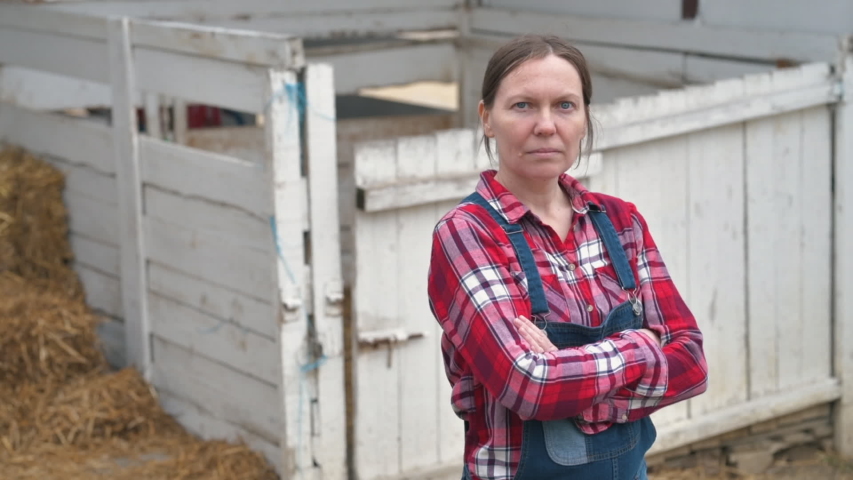 Serious female farmer posing on farm. Confident woman farm worker wearing plaid shirt and jeans overalls looking at camera, arms crossed