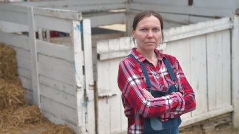 Serious female farmer posing on farm. Confident woman farm worker wearing plaid shirt and jeans overalls looking at camera, arms crossed