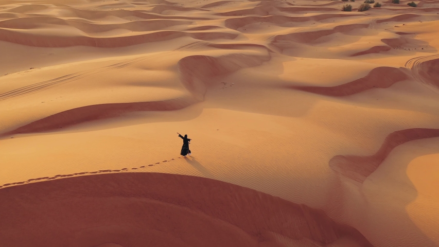Aerial view from a drone flying next to a woman in abaya United Arab Emirates traditional dress walking on the dunes in the desert of the Empty Quarter. Abu Dhabi, UAE.