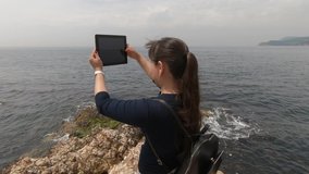 A tourist with a backpack behind shoots a video of sea waves on his tablet