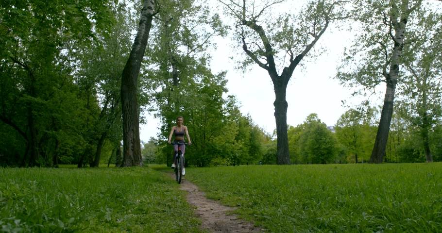 Beautiful slender girl in a tank top and leggings riding a bike among the trees and green grass.