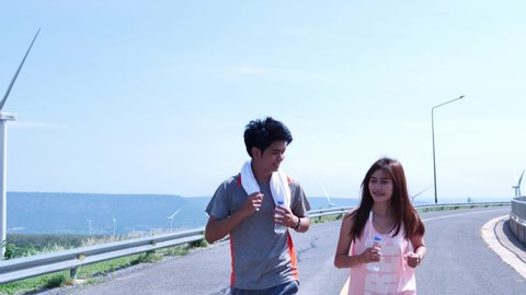 Young Asian couples jogging and hold drinking water bottle on the street in the morning. Wind turbines in background. Runing exercise, Health care lifestyle. Slow Motion 스톡 비디오
