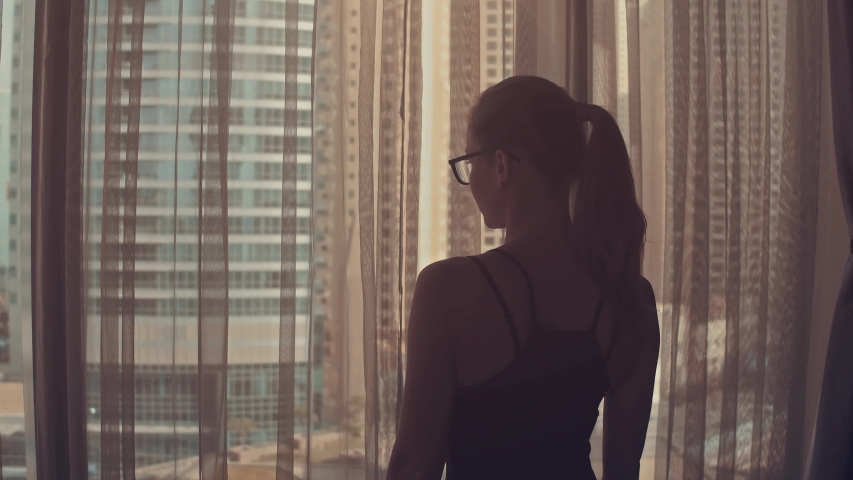 Silhouette of young woman opens curtains on the big window stretches arms and looking out her apartment on the city buildings during amazing sunrise. Dubai city | Shutterstock HD Video #1031095883