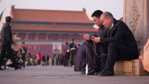BEIJING - MARCH 23, 2018: Two unidentified men sitting at Tiananmen square with smartphones, people stroll around, Gate of Heavenly Peace building seen on background.