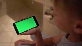 Closeup view of smartphone with green empty blank touch screen in hand of little kid sitting in toilet and watching something using modern mobile device.