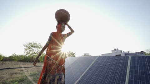 Indian woman wearing sari  and carrying water jug, walking past solar panels installed in desert area. Rajasthan. India.