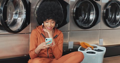 Smiling young woman using smartphone at laundromat. African American student having good news on smartphone. Self-service public laundry.