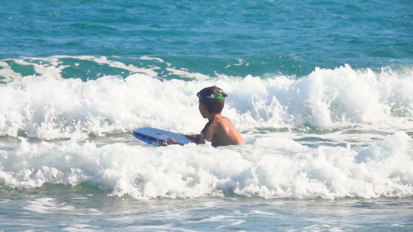 Lttle boy is jumping in the waves on a small surfboard. Summer sunny day at the sea.