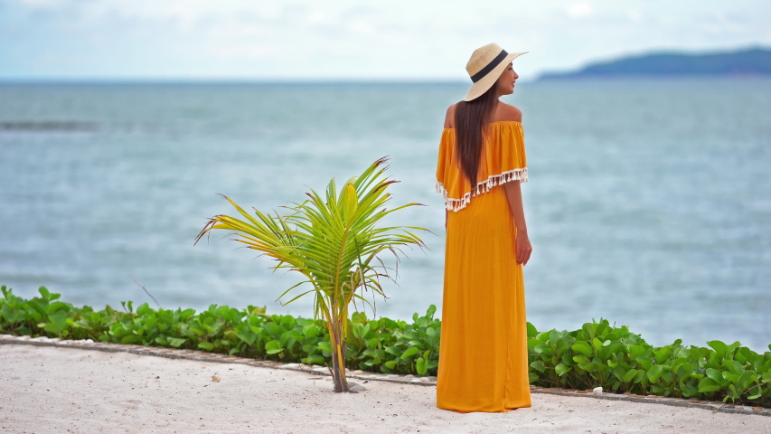 A beautiful girl standing in an orange summer dress, looks relaxed at the sea in a vacation setting