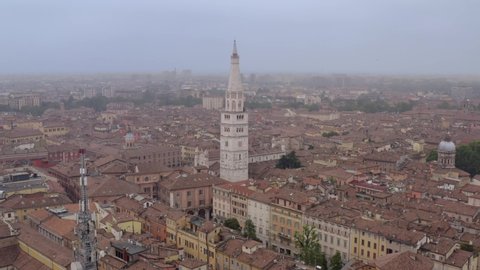Forward drone shot of the Ghirlandina tower, Modena, Italy, in the middle of the city, surrounded by buildings and streets, on a cloudy and foggy day