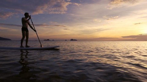 silhouette of man on SUP paddle standing board at sunset, beach leisure activity video