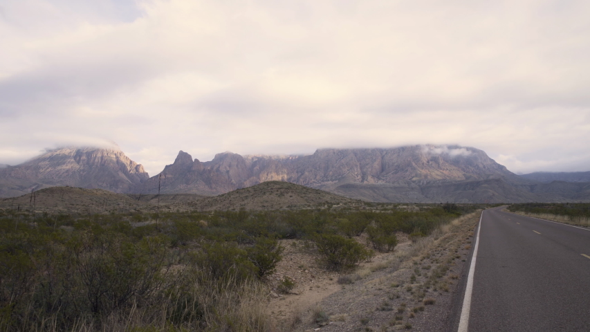 Pull back wide shot of tall mountain range covered by clouds, asphalt road cutting through the scenery