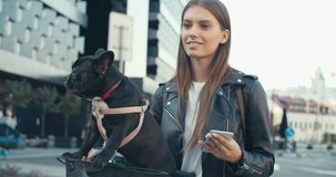 Smiling young woman and black French bulldog walking at city street . Girl with electric kick scooter. 4K slow motion raw video footage 60 fps