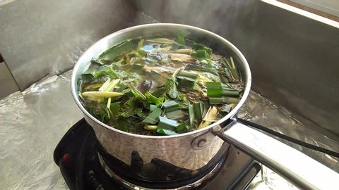Boiling Thai herbs which are sliced lemon grass,  pandan leaves and mint leaves together with white smoke floating above the stainless steel cooking pot