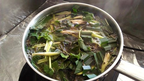 Closeup boiling Thai herbs; sliced lemongrass, pandan leaves and mint leaves; in a stainless steel pot with white smoke floating above