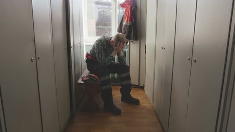 Unhappy caucasian engineer in uniform has difficulties with his work, sits in locker room at construction site, clutches his head in despair, closes face with hands