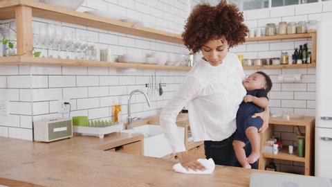 Multi-Tasking Mother Holds Sleeping Baby Son Whilst Cleaning And Working On Laptop In Kitchen