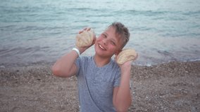 Closeup view of cute happy smiling white kid playing outside with two seashells while sitting at beach of resort. Slow motion full hd video footage.