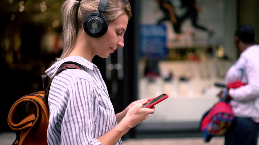 Slow motion of young woman in casual wear walking on street and chatting online on smartphone using public internet.Hipster girl with backpack listening music in headphones and strolling in city Royalty-Free Stock Footage #1031172707