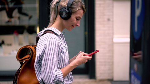 Slow motion of young woman in casual wear walking on street and chatting online on smartphone using public internet.Hipster girl with backpack listening music in headphones and strolling in city