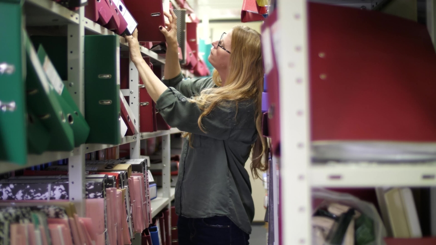 Blonde woman in glasses looking for the binder on the shelves of archive Royalty-Free Stock Footage #1031173226