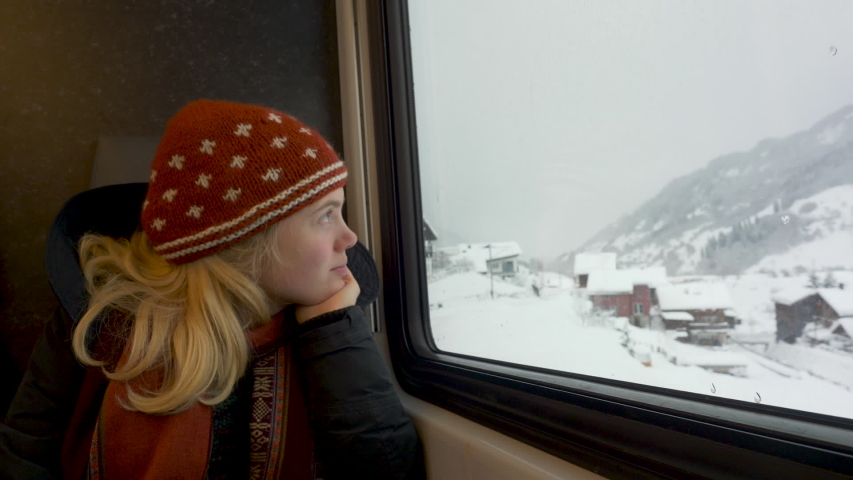 A woman train passenger looking out of window at the snow covered trees, wearing a red hat and scarf