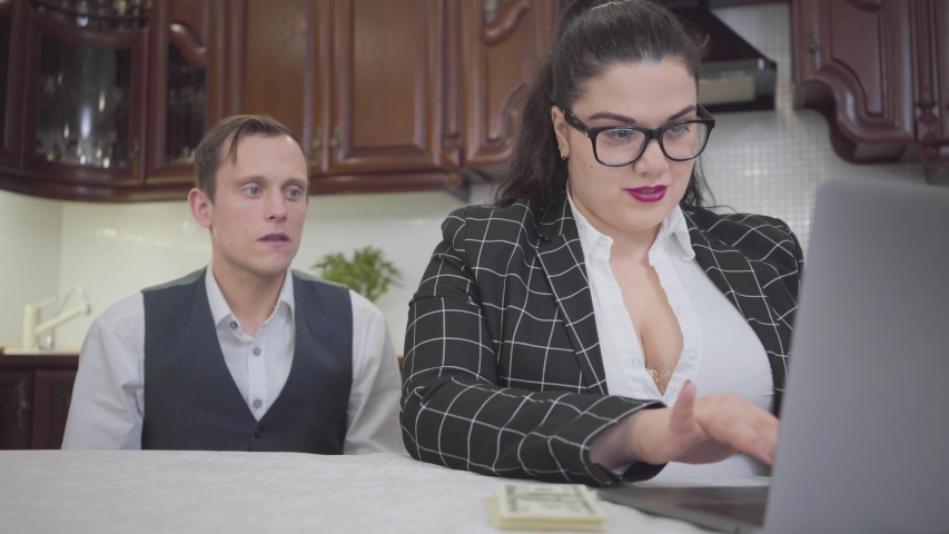 Portrait cute young confident plump woman in glasses working with laptop at the table. Timid young man sitting near wife. Lady boss gives money to husband. Family relationship.