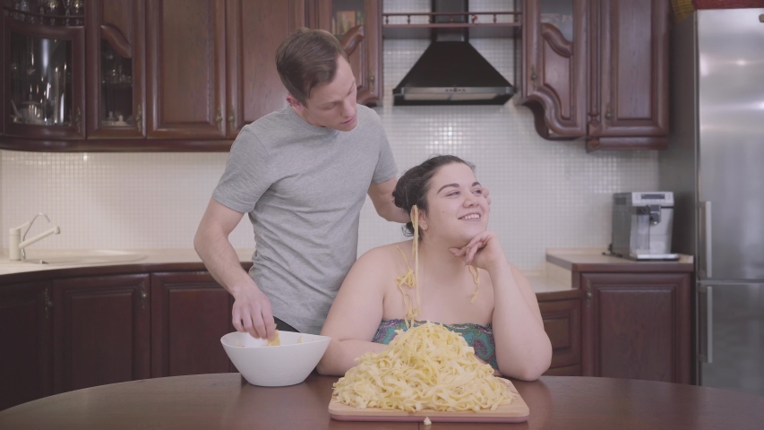 Portrait of adorable plump woman sitting in the kitchen at home. The slim blond man hanging noodles on the ears of his fat girlfriend. False relationships of obese lady and handsome man