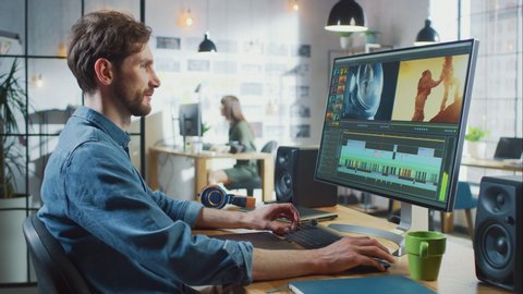 Male Video Editor with Beard and Jeans Shirt Works with Footage on His Personal Computer with Big Display. He Works in a Cool Office Loft. Other Female Creative Colleague Walks in the Background.