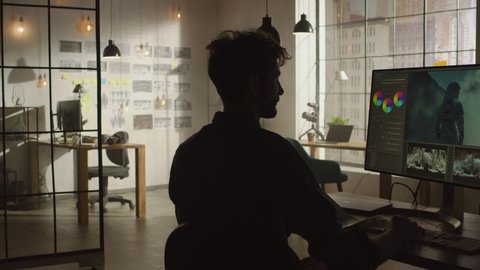 Young Creative Man Stands Up from His Desk with Personal Computer and Display with Video Editing Software and Comes to the Wall with Organized Storyboard in His Cool Loft Office. Sunny Day.