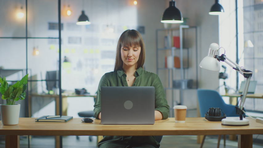 Young Beautiful Brunette Works on a Laptop Computer in Cool Creative Agency in a Loft Office. She has a Take-away Coffee and a Notebook on Her Table. Her Colleague Walks in the Background. Royalty-Free Stock Footage #1031177369