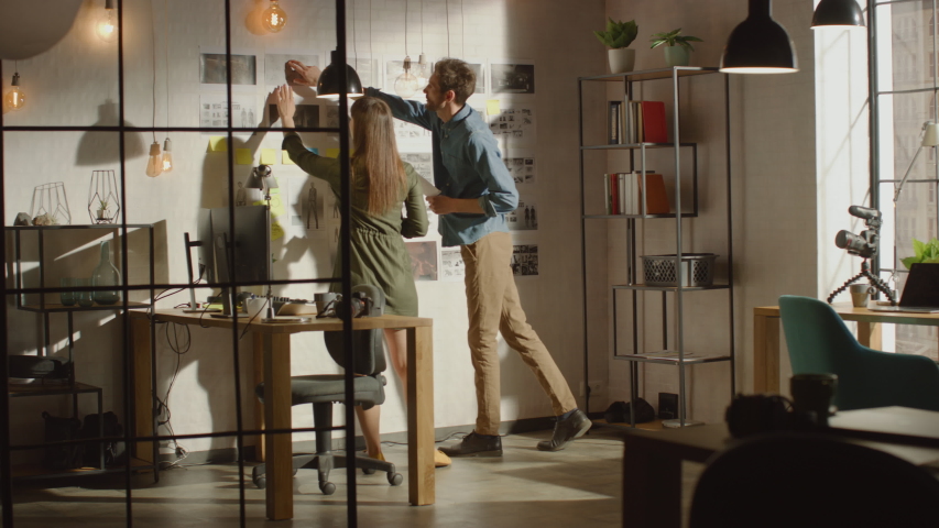 Young Creative Male and Female are Organizing a Mood Board on a Wall of Their Cool Office Loft. Pictures Represent Future Motion Video. Colleagues are Working and Discussing the Project. Sunny Day. Royalty-Free Stock Footage #1031177393