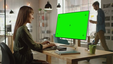 Beautiful Professional Creative Employee Works on Her Personal Computer with Big Green Screen Mock Up Display. She Works in a Cool Office Loft. Other Male Colleague Walks in the Background.