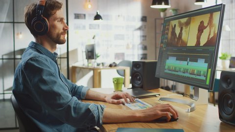 Male Video Editor Puts Headphones and Starts Working with Footage on His Personal Computer with Big Display. He Works in a Cool Office Loft. Creative Man Wears a Jeans Shirt.