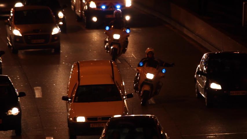 Paris, France - April 6, 2019: Motorcycle policemen and Ambulance with emergency lights flashing in Car Traffic by night at Paris. Lights, cars, fires, bus, blur effect. Lot of vehicles. Close up.