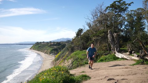A strong man running in slow motion on a nature trail on the edge of an ocean cliff with epic views of the beach and California coastline in Santa Barbara.