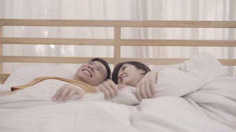 Asian couple lying on bed in bedroom, couple feeling happy having funny time playing under blanket together on bed at home. Couple relax enjoy love moment at home concept.