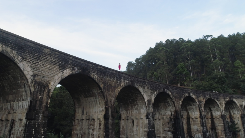 Low angle pan, woman in red walks alone across tall Nine Arch Bridge in Ella, Sri Lanka. Forest far below